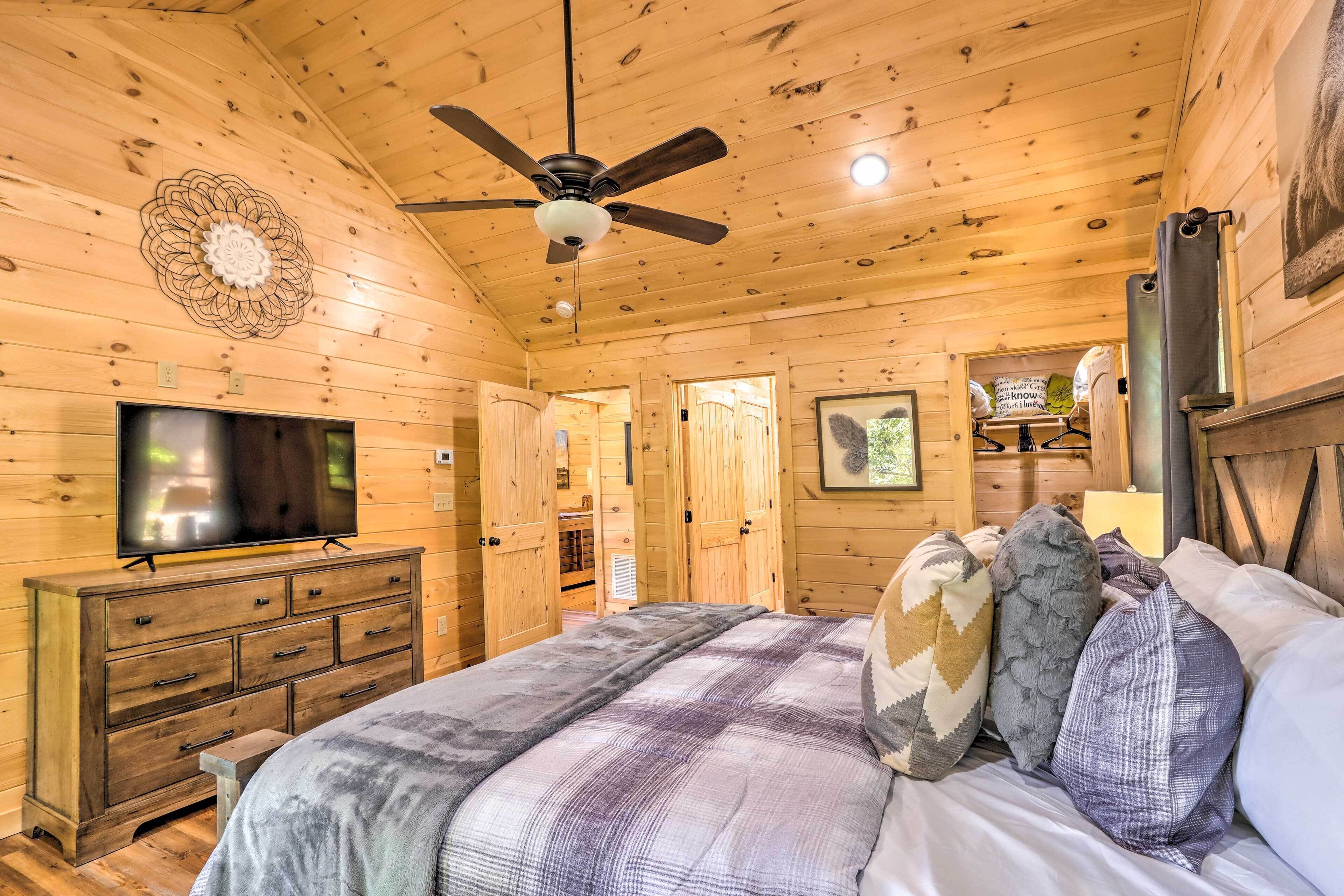 A wooden cabin bedroom with a bed, pillows, a dresser, a television, and a ceiling fan.