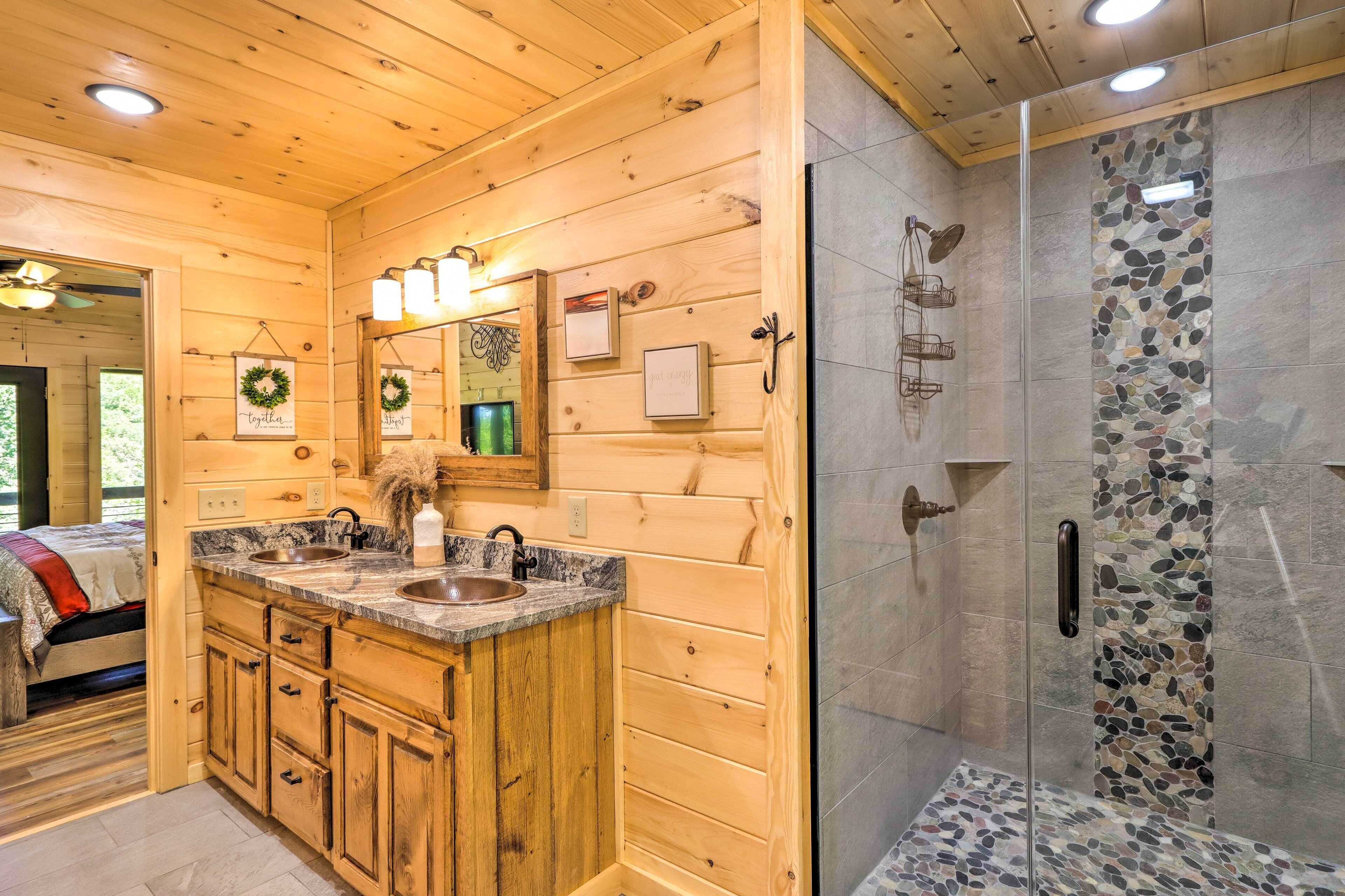 Ein Badezimmer einer Holzhütte mit einem doppelten Waschbecken, einer Dusche mit Steinwand und einem sichtbaren Bett im Hintergrund.