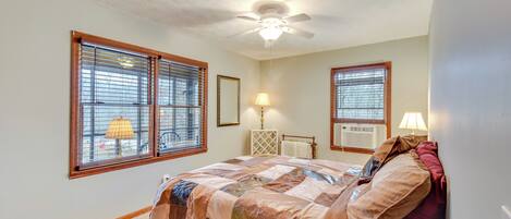 A bedroom with a bed, two windows with blinds, a ceiling fan, and a nightstand with a lamp.