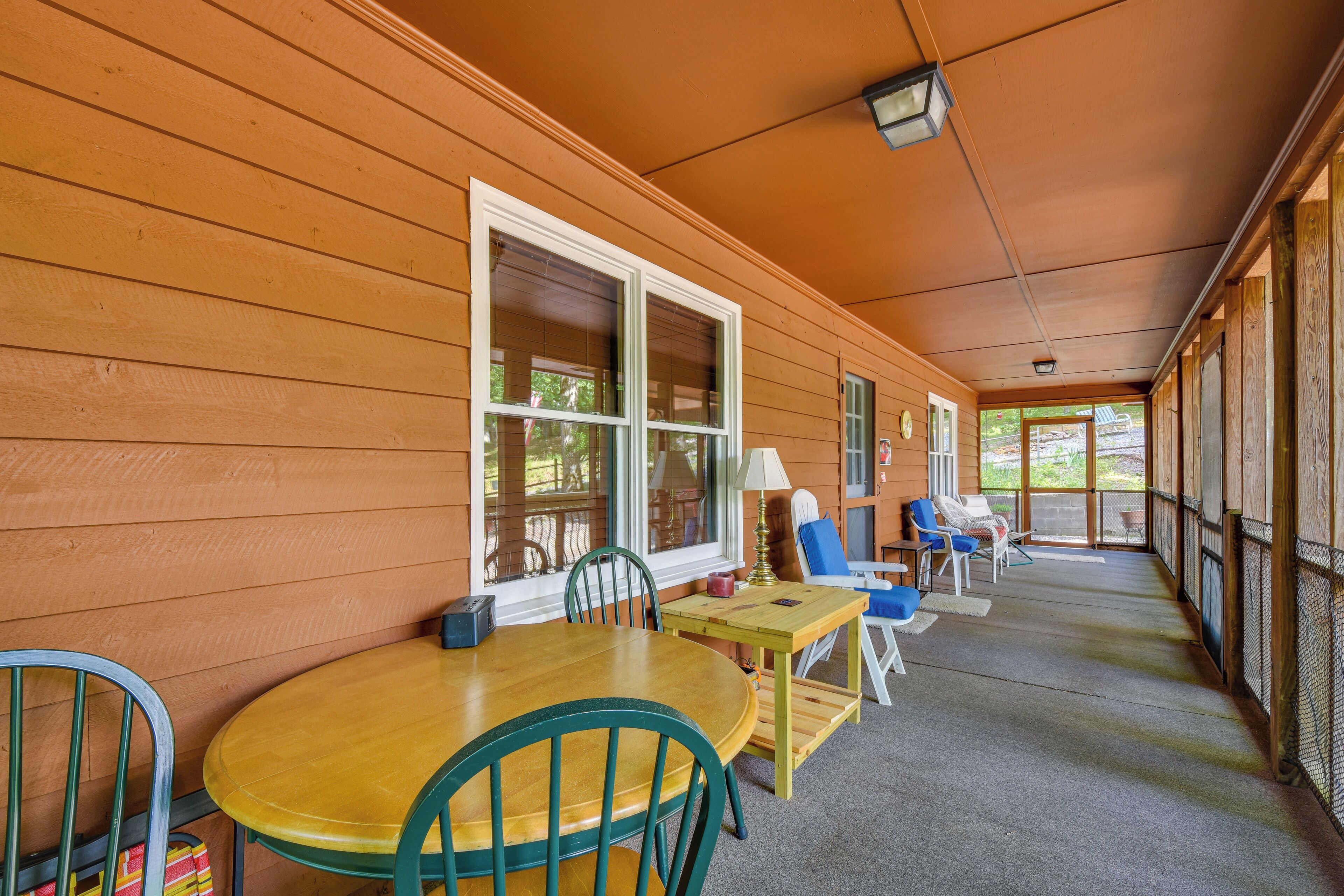 A covered porch with a wooden table and chairs, a small table with a lamp, and a view of the outdoors.