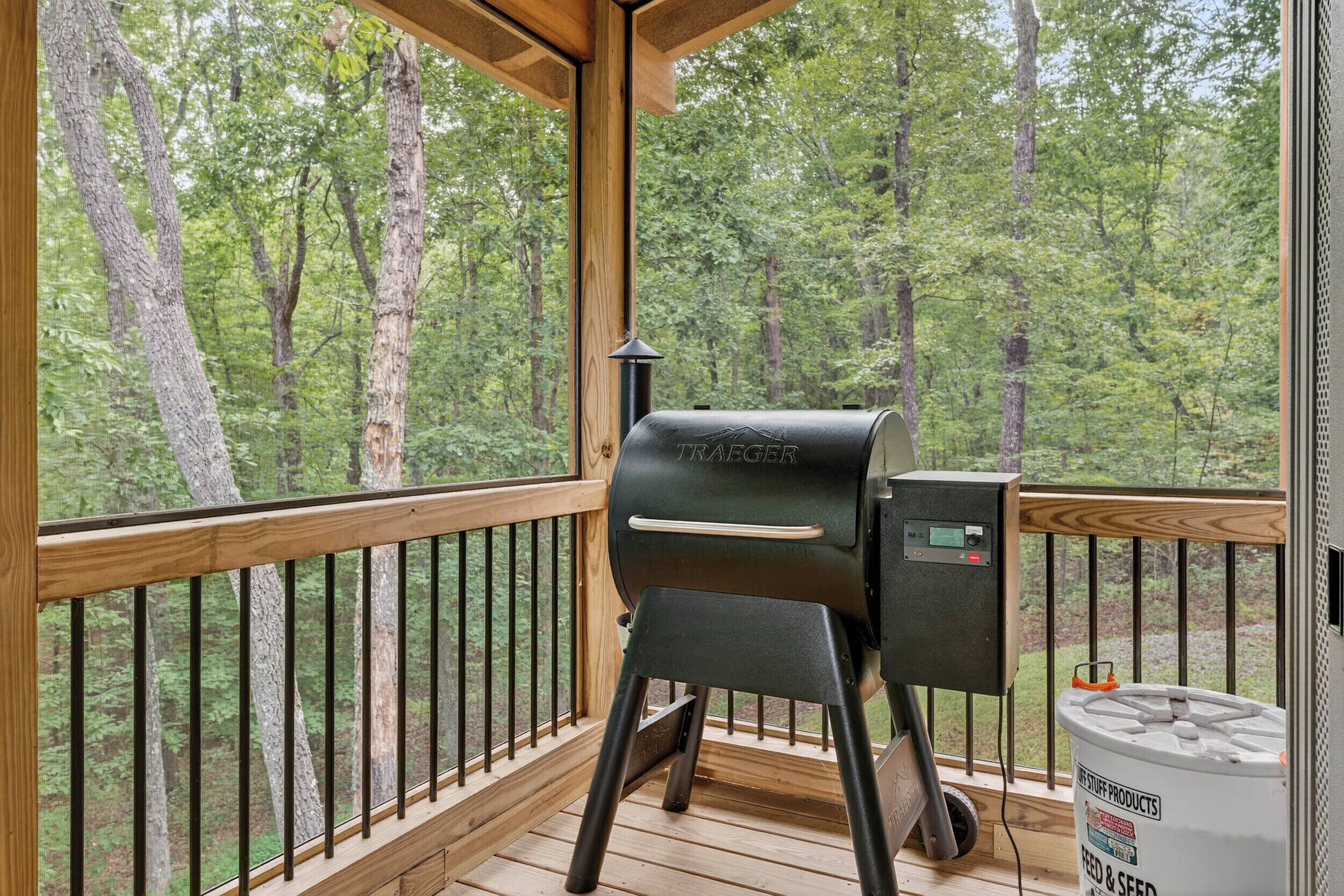 Une terrasse en bois avec un barbecue et un récipient portant l’étiquette « PRODUITS POUR L’ALIMENTATION ET LE SEMIS », le tout situé sur une véranda donnant sur une forêt.