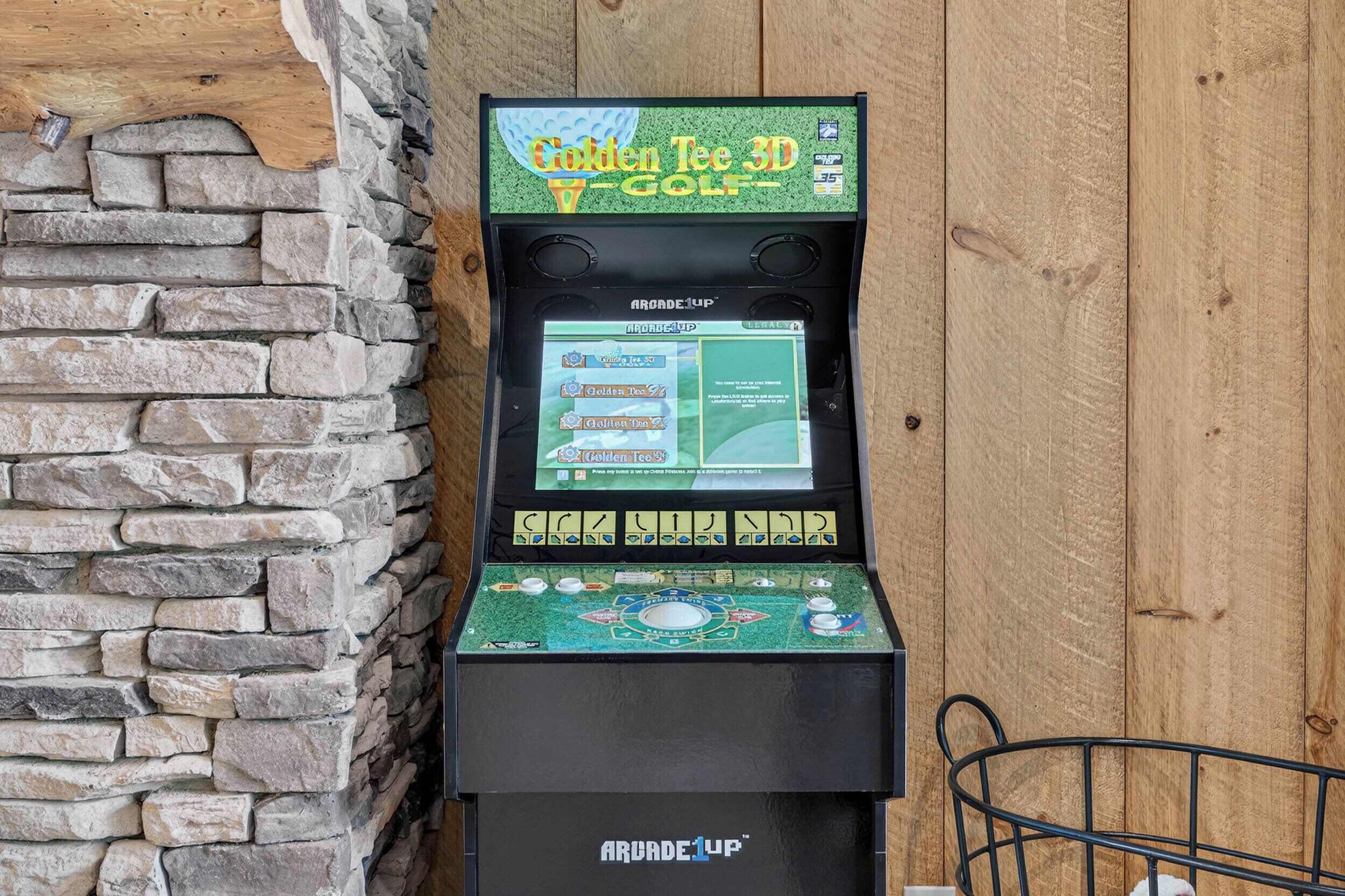 Máquina de arcade 3D para golfe com a marca 'Golden Tee 3D Golf' pela ARCADEUP, posicionada em frente a uma parede de pedra com uma cadeira de metal por perto.