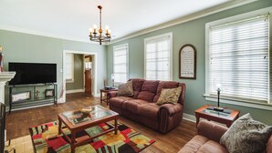 A living room with a brown leather sofa, a glass coffee table, and a colorful rug. There is a television on a stand, a chandelier, and a window with blinds. - Cozy Jackson Vacation Rental < 1 Mi to University (Jackson)