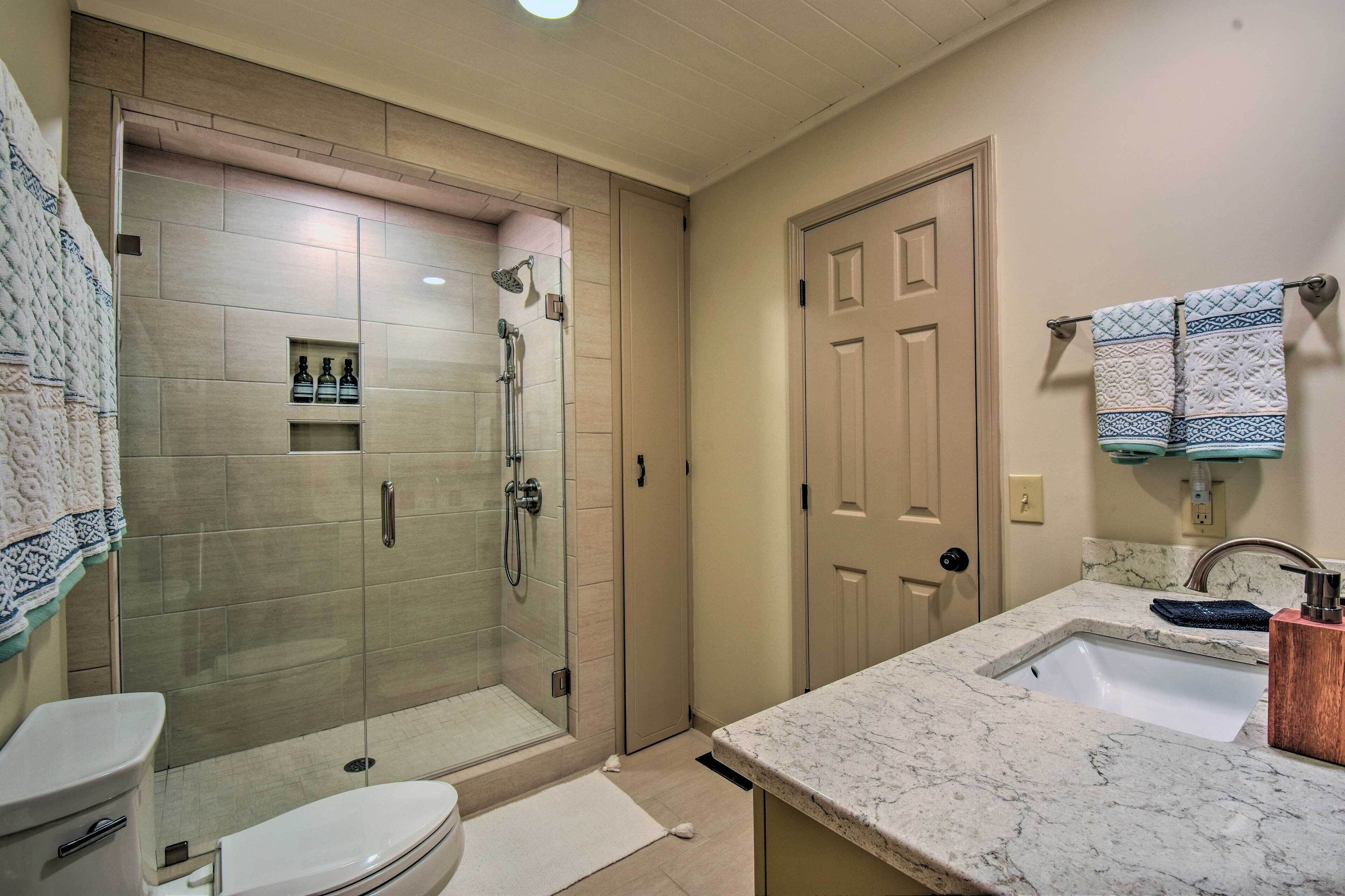 Ein Badezimmer mit einer gläsernen Duschkabine, einer Toilette, einem Waschbecken mit Marmorplatte und einem Handtuchhalter mit gemusterten Handtüchern.