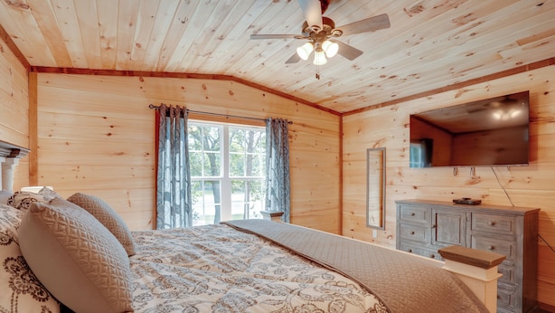 Un dormitorio de cabaña de madera con cama, cortinas, ventilador de techo, televisor y cómoda.