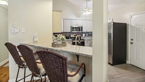 A kitchen with white cabinets, a stainless steel refrigerator, and a microwave. There is a dining area with wicker chairs and a granite countertop. - Savannah Home w/ Patio & Grill ~ 2 Mi to Downtown (Savannah)