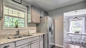 Una cocina con armarios blancos, un refrigerador de acero inoxidable, un lavamanos doble y una ventana con una planta.