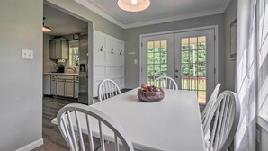 Un comedor con una mesa blanca, sillas y un frutero. Se observa una cocina de fondo con muebles blancos y un refrigerador.