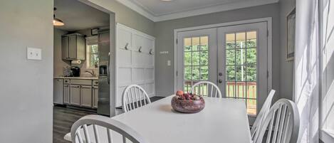 A dining area with a white table, chairs, and a fruit bowl. There is a kitchen in the background with white cabinets and a refrigerator.