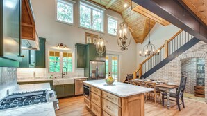 A spacious kitchen with wooden cabinets, a marble countertop, and a central island. There is a dining area with a brick fireplace and a staircase leading to an upper level. - Luxurious Clarkesville Cabin w/ Decks & Fire Pit (Clarkesville)