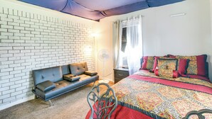 A bedroom with a brick wall, a sofa, a bed with patterned bedding, a standing fan, and a window with curtains. - Horn Lake Home w/ Yard: 8 Mi to Graceland! (Horn Lake)
