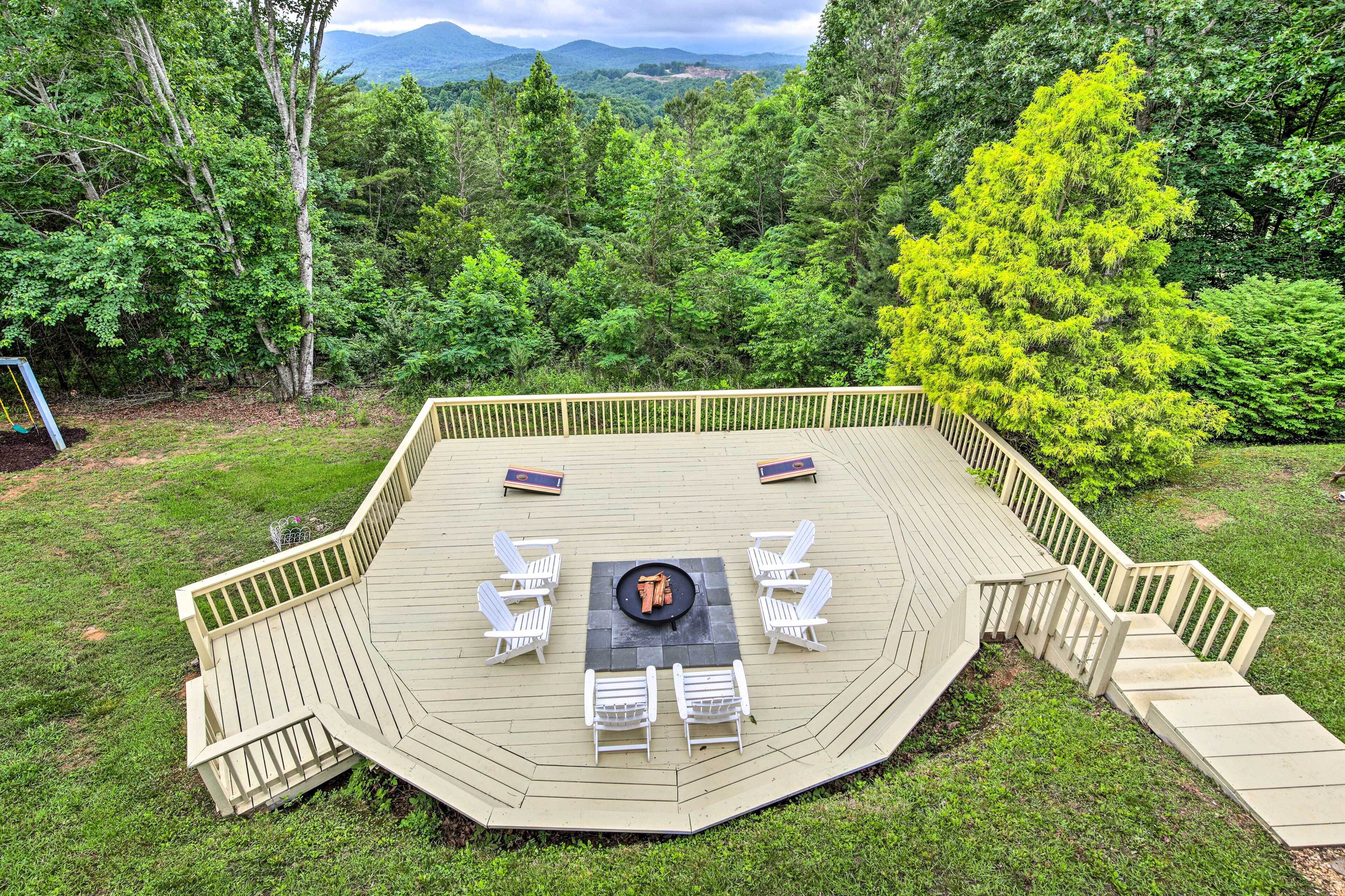 A deck with a fire pit, white chairs, and a view of a forest and mountains.