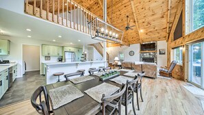 Un intérieur spacieux avec un plafond en bois, un coin repas doté d’une grande table et de chaises, une cuisine avec des meubles de couleur vert clair et un coin salon confortable agrémenté d’une cheminée.