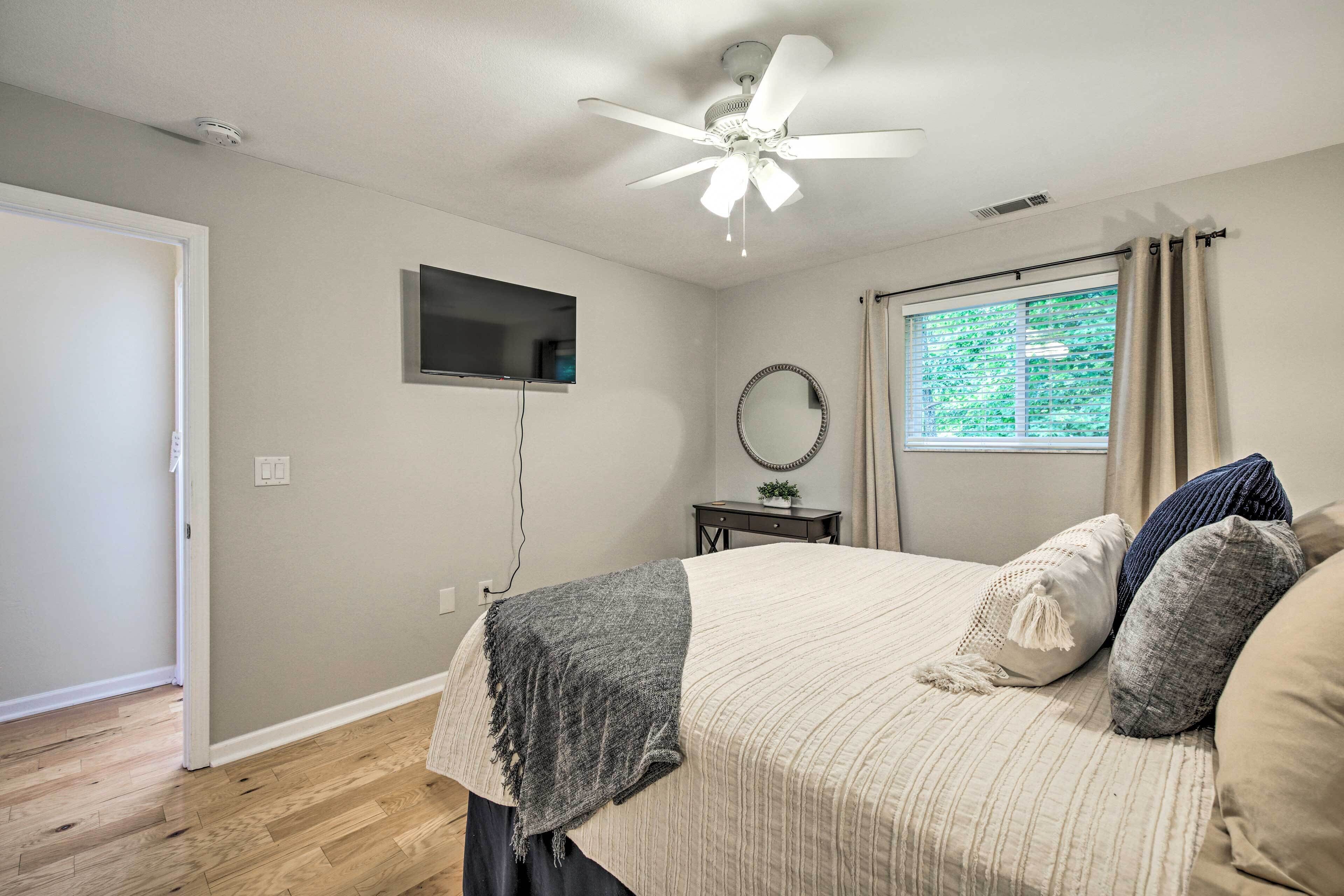 A bedroom with a bed, a ceiling fan, a TV, a window with curtains, a bedside table, and a mirror.