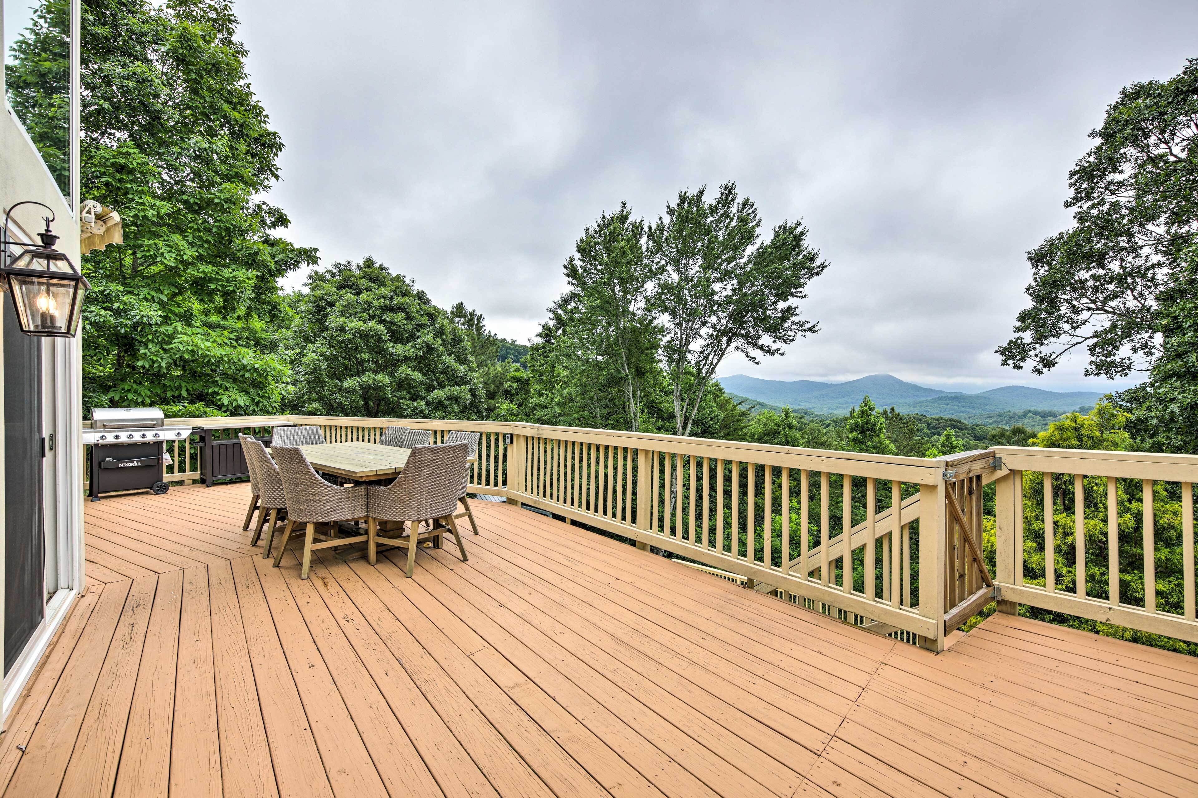 A wooden deck with a table and chairs, a barbecue grill, and a view of mountains and trees.