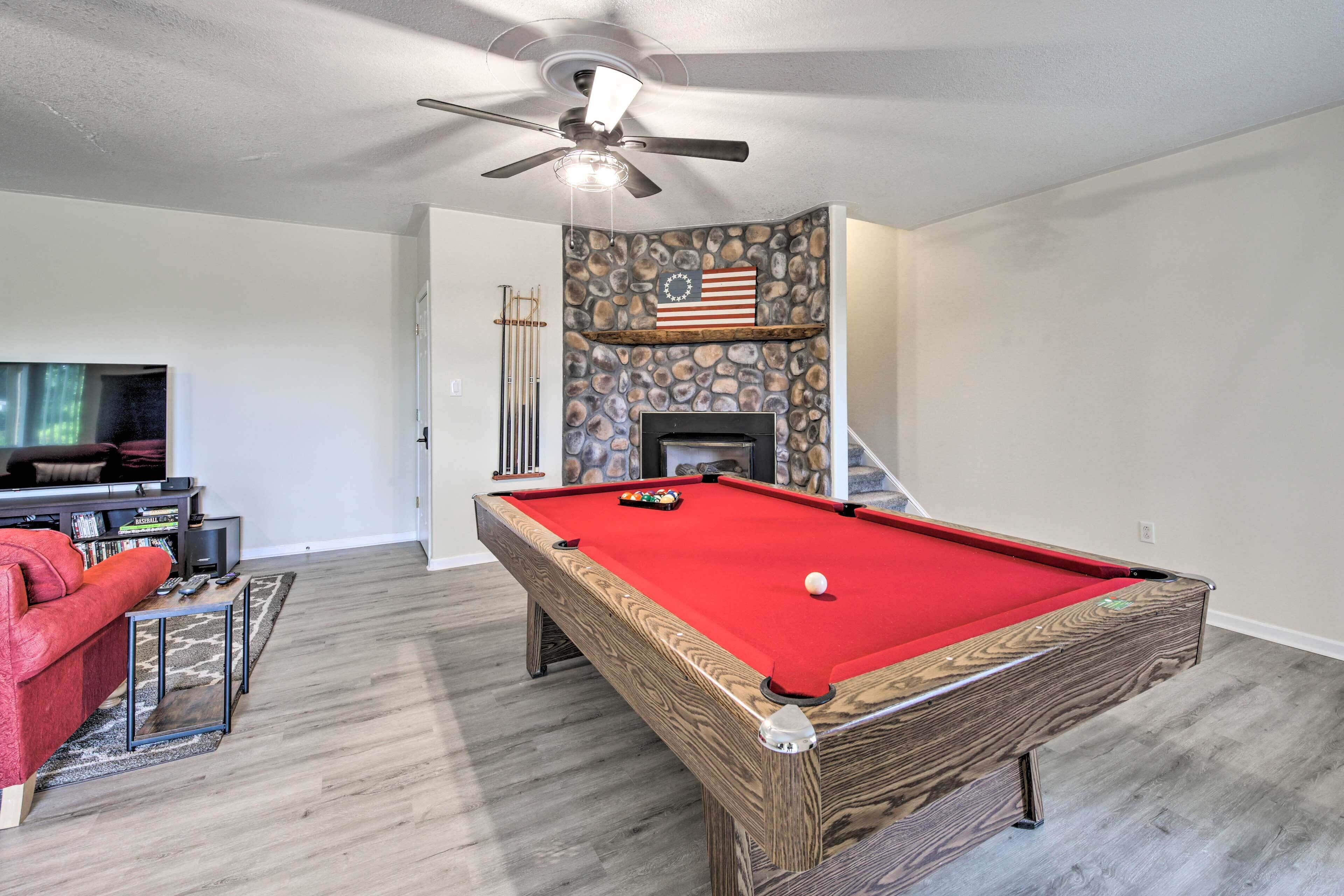 A pool table with a red felt surface, a ceiling fan, a stone wall with an American flag, and a fireplace.