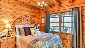 Ein Schlafzimmer einer Holzhütte mit einem Bett, Nachttischen, einer Chandelierleuchtre und einem Fenster mit Vorhängen.
