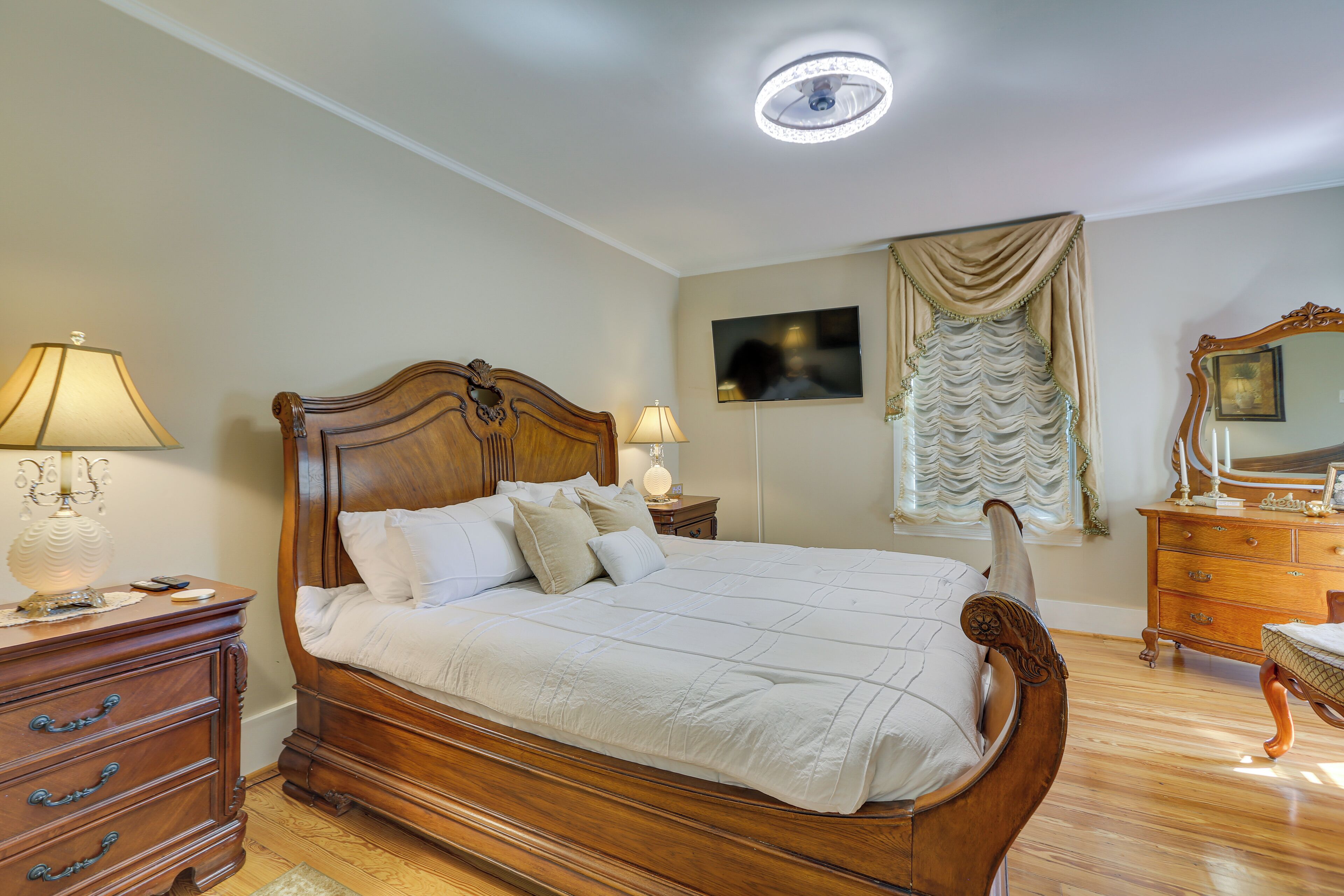 A bedroom with a wooden bed, two bedside tables, a dresser, a television, and a ceiling light.