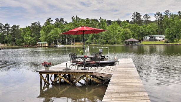 A wooden dock extending into a calm lake, with a table and chairs under a red umbrella, surrounded by lush greenery and a small gazebo.