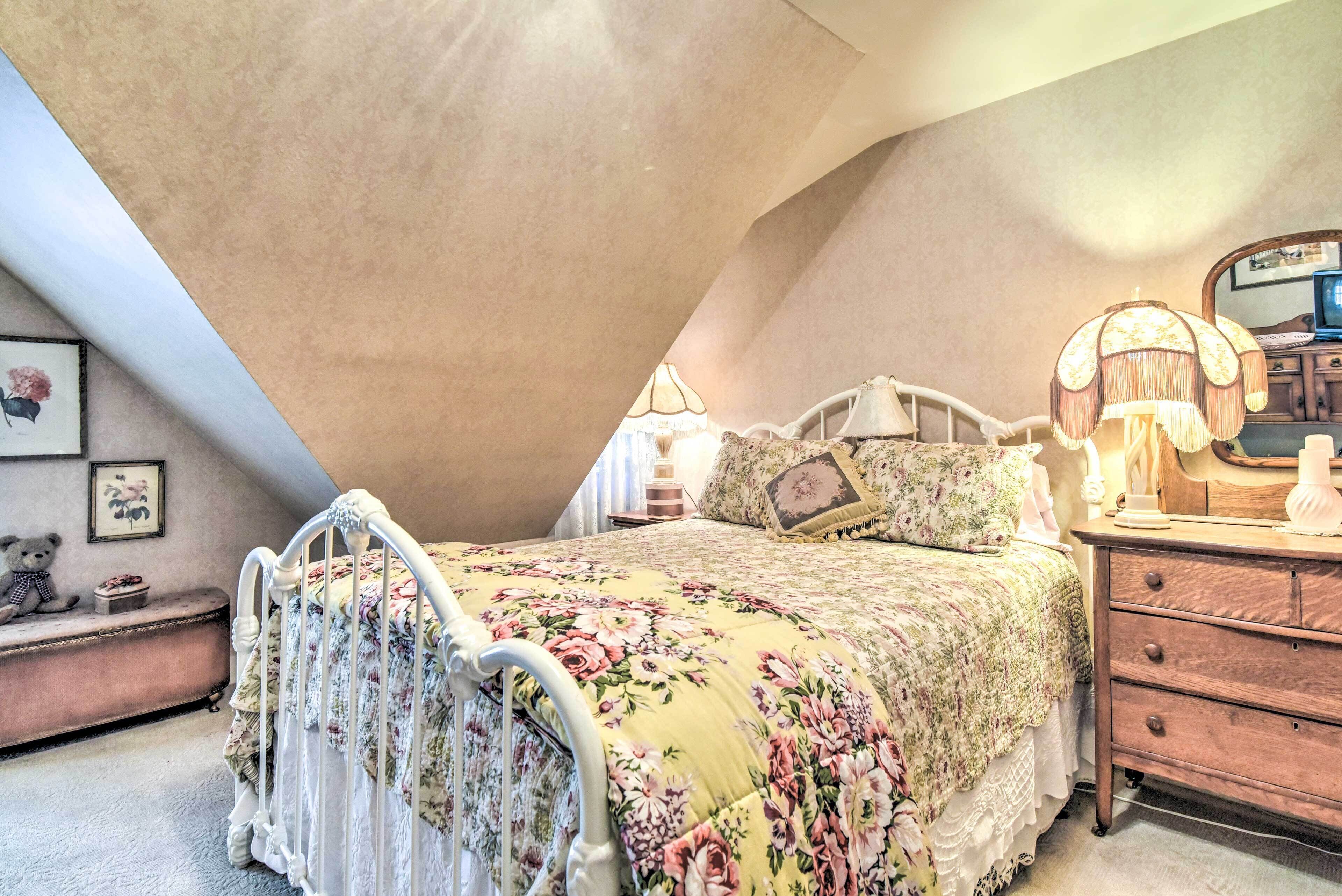 A bedroom with a sloped ceiling, a floral bedspread, a vintage-style bed, a wooden dresser, and a bedside lamp.