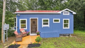 A small blue house with a red roof, a porch with wicker furniture, and a wooden door. - Pendergrass Tiny Home Cabin on Pond w/ Fire Pit! (Pendergrass)