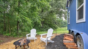 A blue building with white windows, surrounded by trees. There are two white Adirondack chairs and a round table with logs. - Pendergrass Tiny Home Cabin on Pond w/ Fire Pit! (Pendergrass)