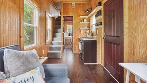 A cozy cabin interior with a wooden interior, a staircase, a kitchen area, and a seating area with a couch and pillows. - Pendergrass Tiny Home Cabin on Pond w/ Fire Pit! (Pendergrass)