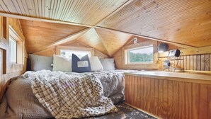 A cozy attic bedroom with a sloped ceiling, a bed with a knitted blanket, a pillow with "DREAM" text, and a small table with a fan. - Pendergrass Tiny Home Cabin on Pond w/ Fire Pit! (Pendergrass)