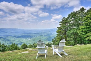Two Adirondack chairs on a grassy hill overlooking a scenic landscape with trees and a view of distant hills.