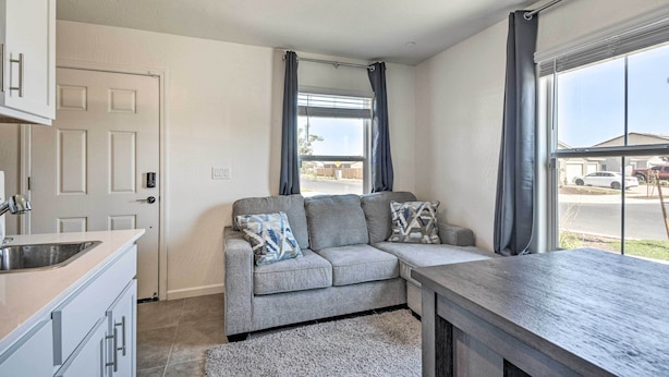 Una sala de estar moderna con un sofá seccional gris, una zona de cocina con pileta y armarios, y ventanas amplias con cortinas.