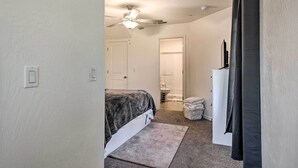 Un dormitorio con cama, ventilador de techo, una puerta blanca y un baño visible al fondo.