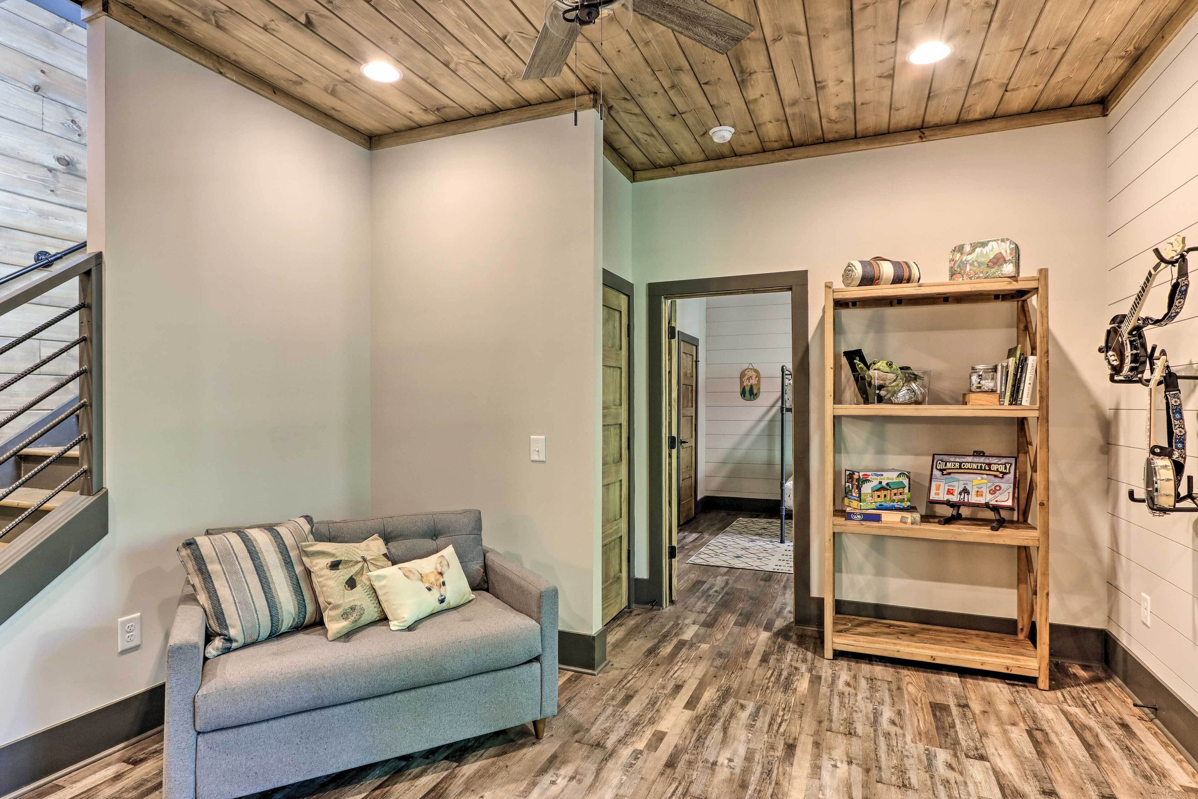 A cozy room with a grey sofa, wooden flooring, a ceiling fan, and a bookshelf with various items.