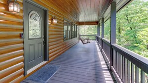 A wooden porch with a dark gray door, a bench, and a railing overlooking a forested area. - Riffle Run Cabin: River Access, Hot Tub, Game Room (Ellijay)
