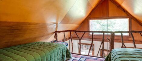 A cozy attic bedroom with two green beds, a wooden ceiling, and a large window.