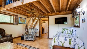 A cozy room with a wooden ceiling, a bed with a deer print duvet, a brown leather sofa, a patterned armchair, and a wooden coffee table. - Cozy Georgia Abode w/ Deck, Fire Pit & Lake Access (Menlo)