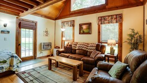 A cozy living room with a wooden ceiling, a brown leather sofa, a wooden coffee table, and a large window with a view of trees. - Cozy Georgia Abode w/ Deck, Fire Pit & Lake Access (Menlo)