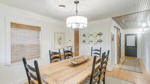 Un coin repas avec une table en bois, des chaises, un lustre et une fenêtre équipée de stores.