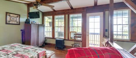 A room with a wooden ceiling, a ceiling fan, a bed with a quilt, a red blanket, a dresser, a television, and large windows.