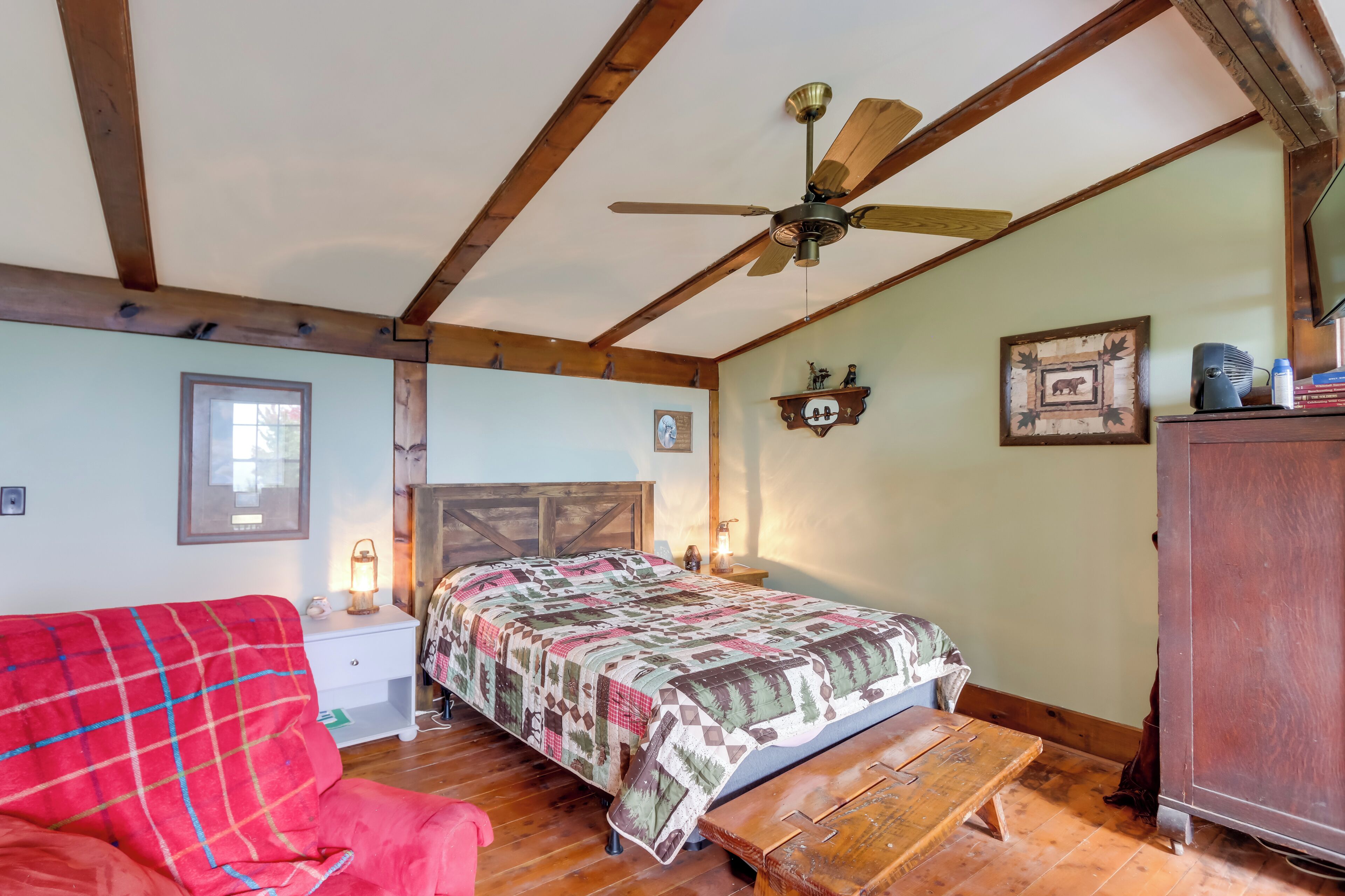 A cozy bedroom with a bed, a wooden bench, a red chair, a ceiling fan, and a dresser.