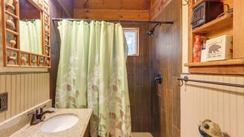 Un baño con tocador de madera, lavamanos, cortina de ducha y estante con artículos de aseo.