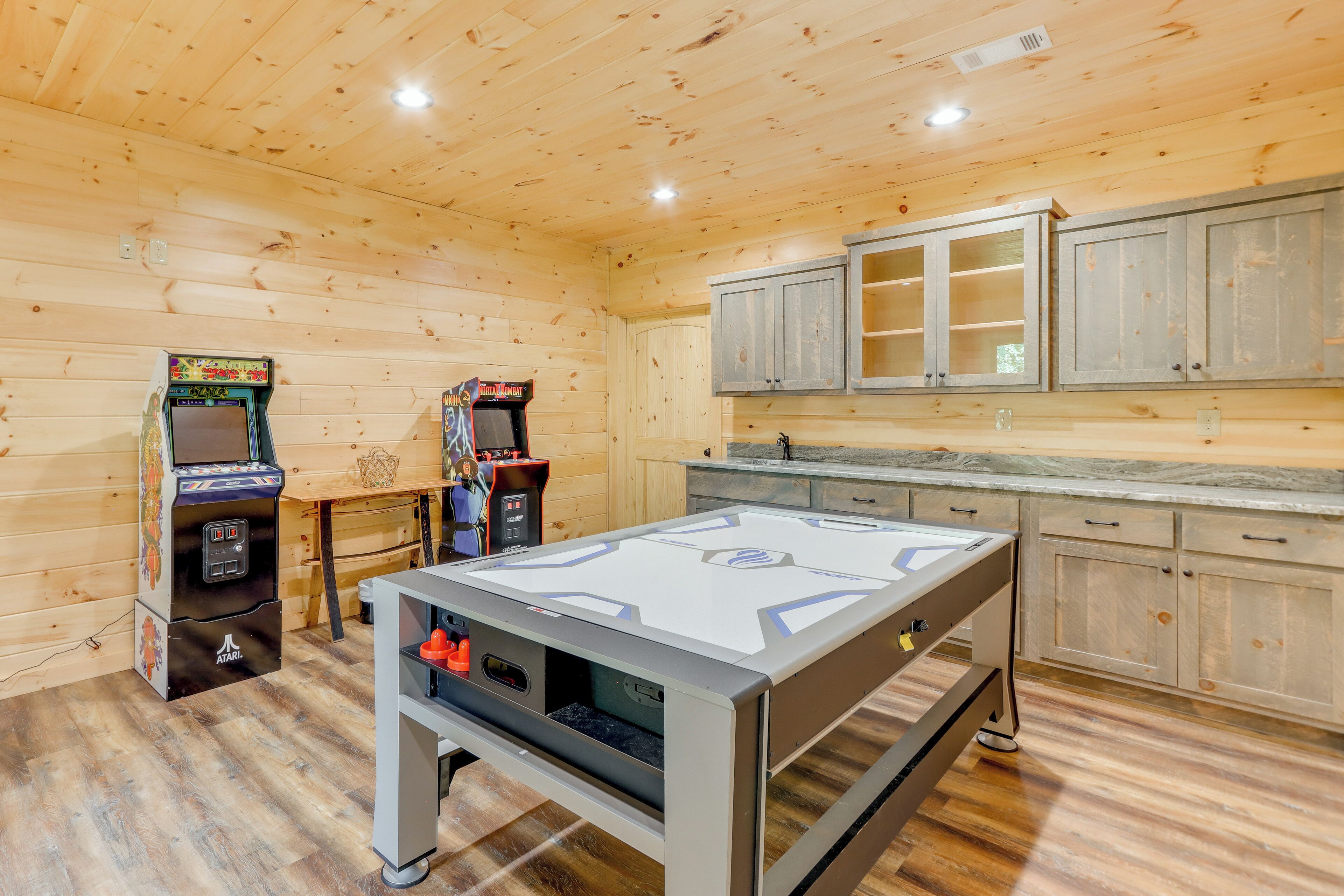 A room with wooden walls and ceiling, a foosball table, two arcade machines, and cabinets.
