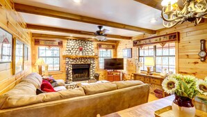 A cozy log cabin interior with a stone fireplace, a large leather sofa, and a television. - Lakefront Blue Ridge Cabin w/ Hot Tub! (Blue Ridge)