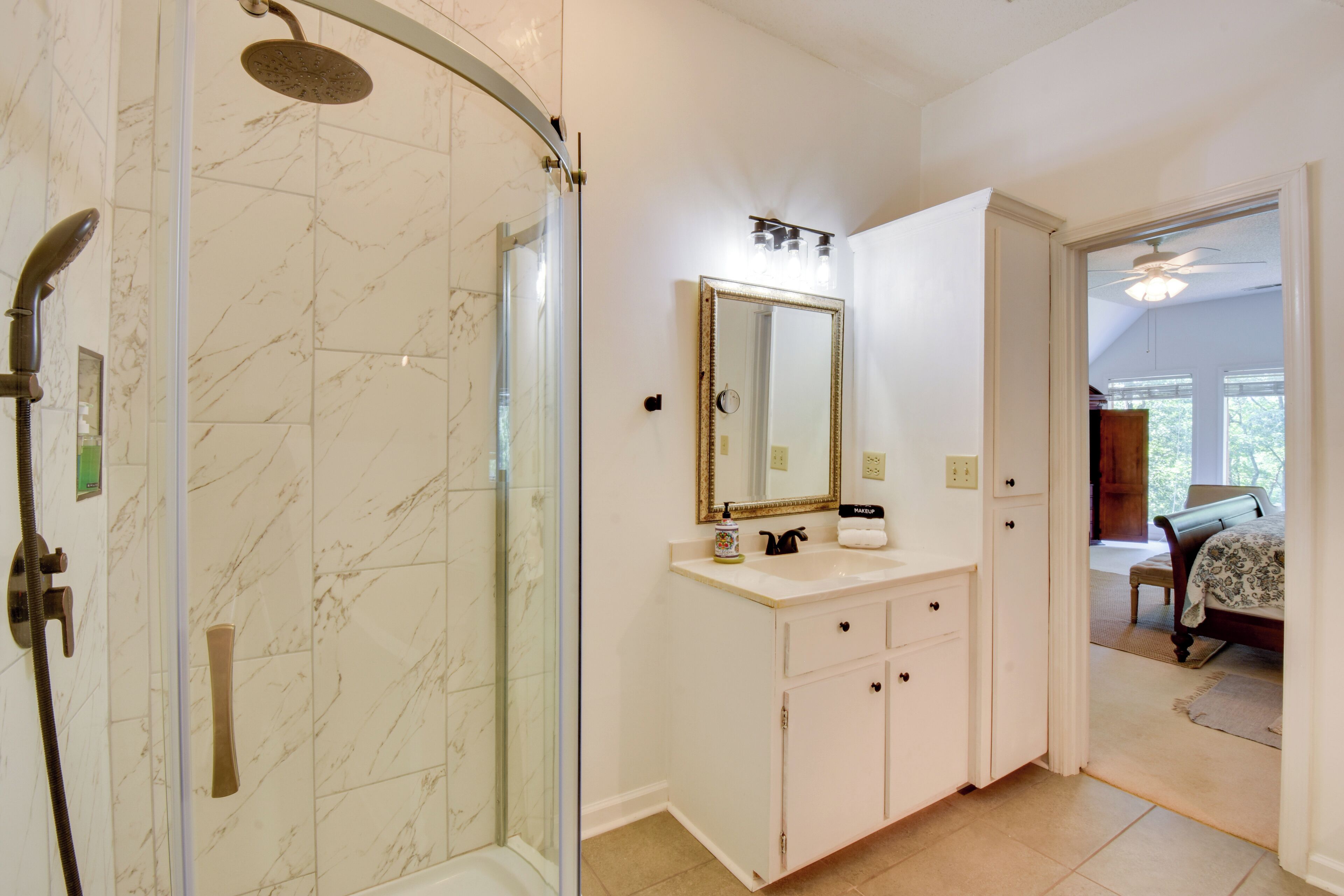 A bathroom with a glass-enclosed shower, a white vanity with a mirror, and a bed visible through an open doorway.