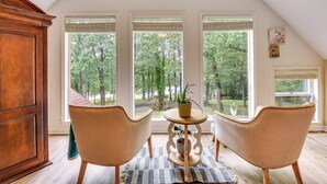 Ein Zimmer mit großem Fenster, einem Holzschrank, zwei Stühlen, einem kleinen Tisch mit Pflanze und einem Teppich.