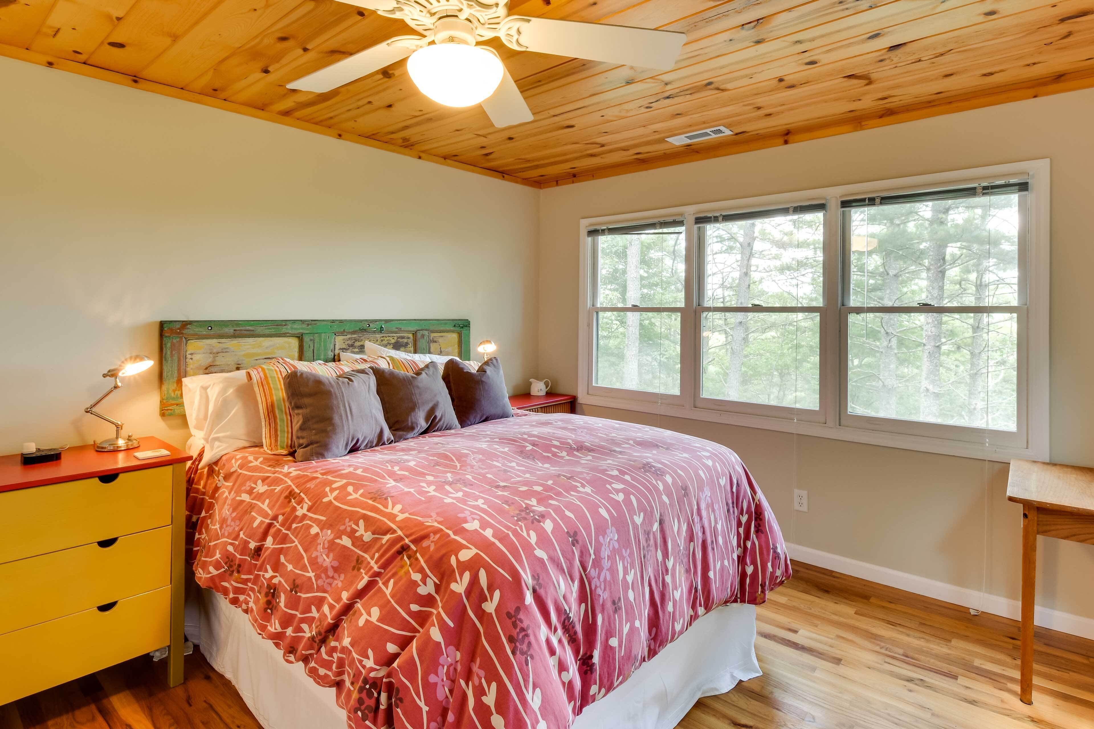 A bedroom with a wooden ceiling, a bed with a floral bedspread, a nightstand with a lamp, and a window with a view of trees.