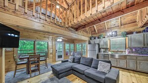 A cozy cabin interior with a large gray sectional sofa, wooden dining table, and chairs. A kitchen area with a refrigerator and cabinets is visible. - 'deer Haven' Cabin: 5 Mi to Dtwn Blue Ridge! (Blue Ridge)
