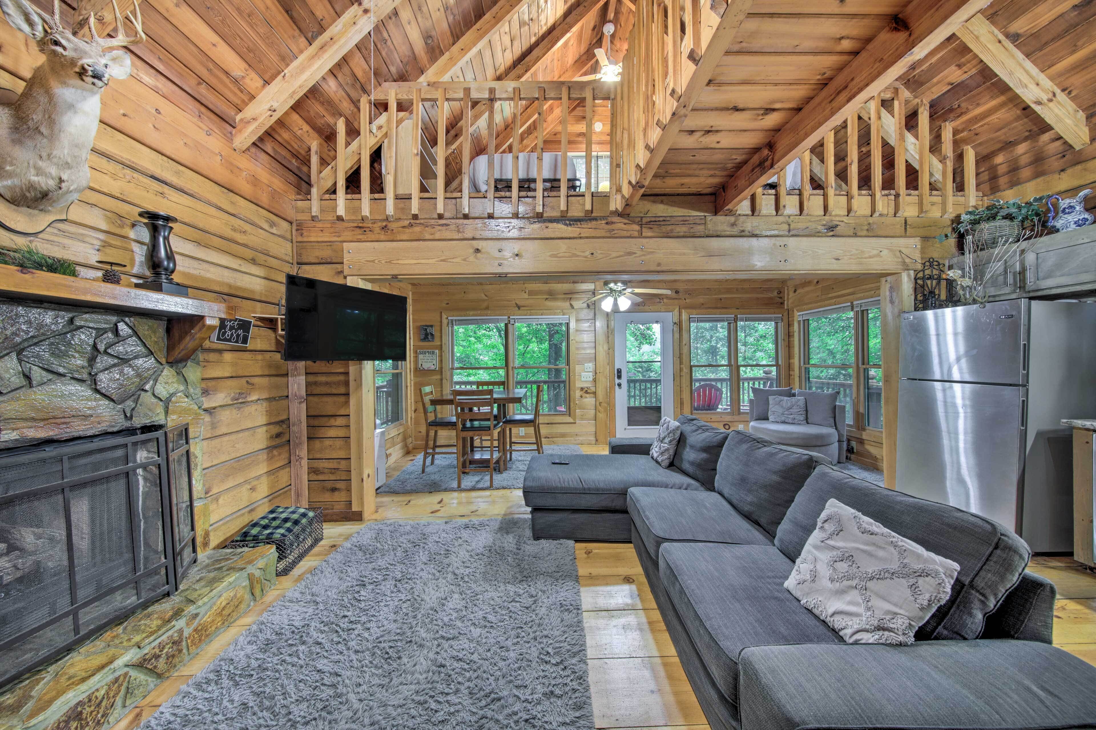 A log cabin interior with a stone fireplace, a gray sectional sofa, a dining area, and a kitchen with a stainless steel refrigerator.