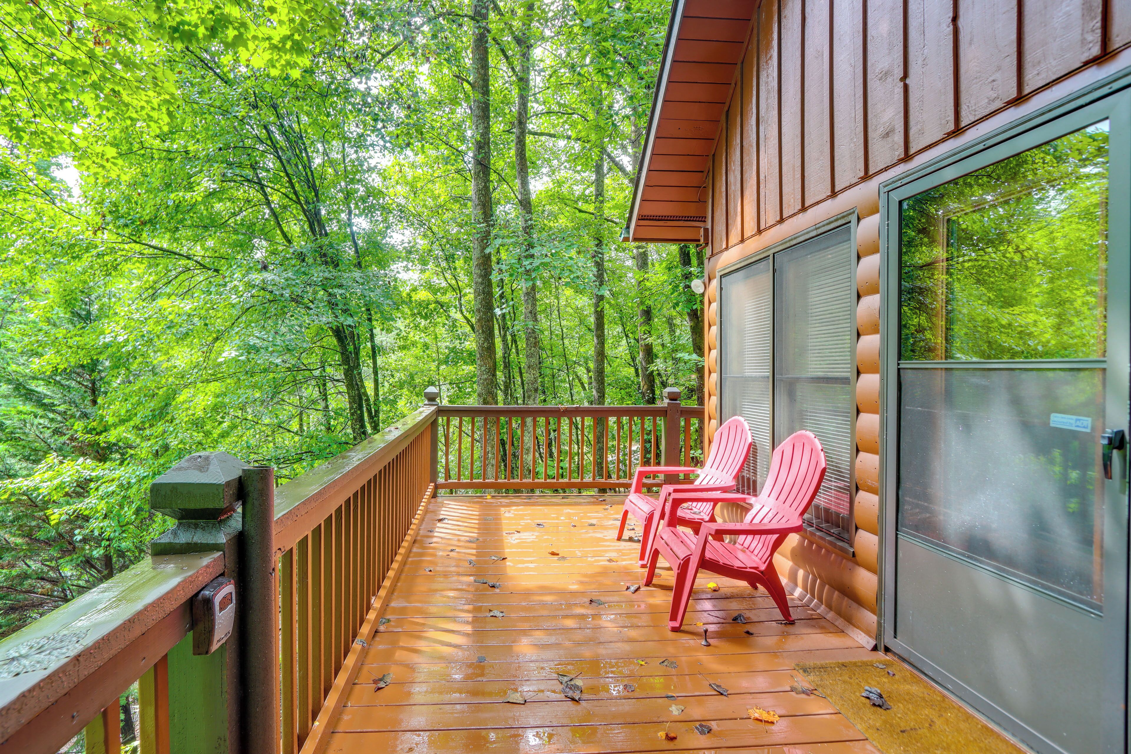 A wooden deck with a railing, two red Adirondack chairs, and a glass door leading to a room.