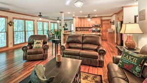 A spacious living room with a leather sofa, a coffee table, and a fireplace. There is a kitchen in the background with wooden cabinets and a refrigerator. - Waterfront West Point Lake Cabin w/ Private Dock! (LaGrange)