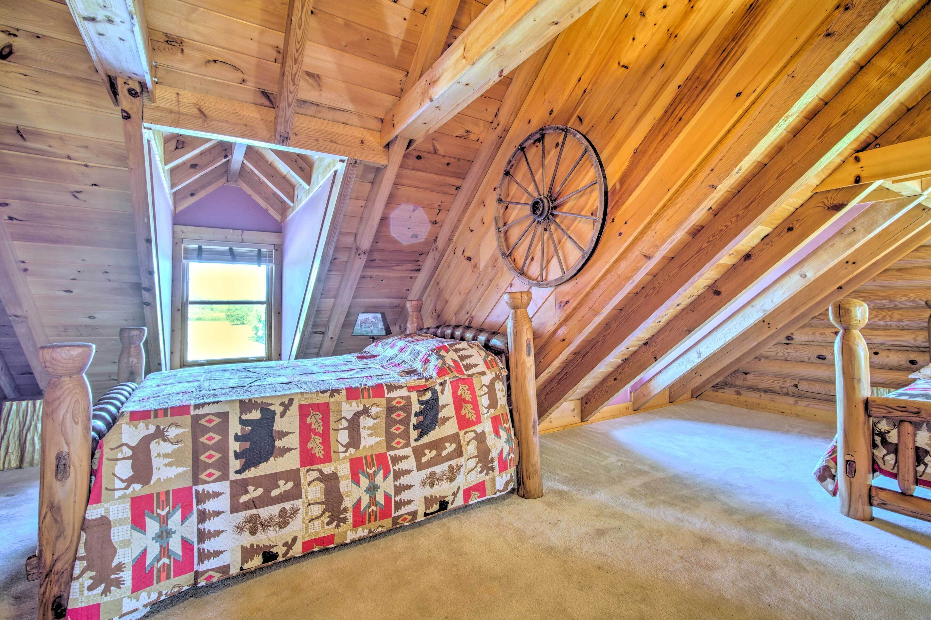 A cozy attic bedroom with a wooden sloped ceiling, a large window, a bed with a patterned quilt, and a wooden wheel on the wall.